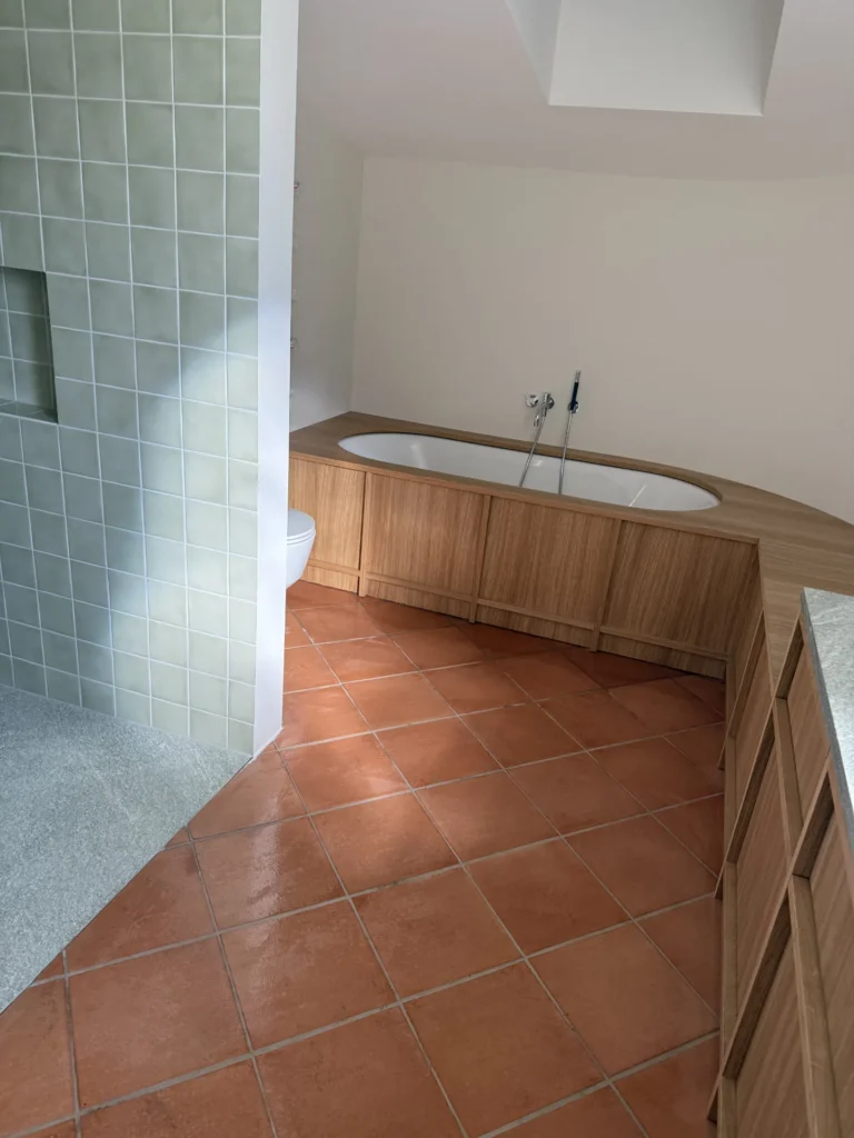Badezimmer mit Terrakotta-Boden, Holzwanne und salbeigrüner Keramik-Duschwand